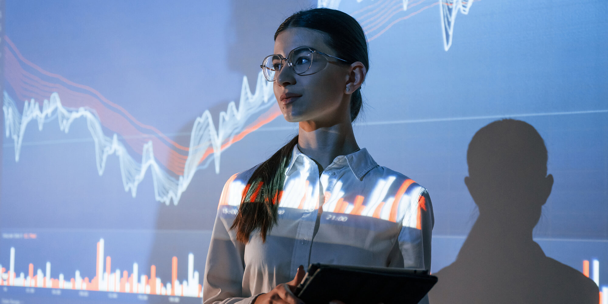 A woman wearing glasses stands before a screen displaying a stock market chart, analyzing financial data
