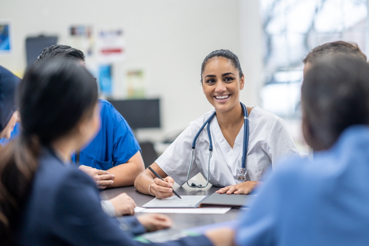 A smiling female nurse sits at a table surrounded by colleagues, fostering a warm and collaborative atmosphere